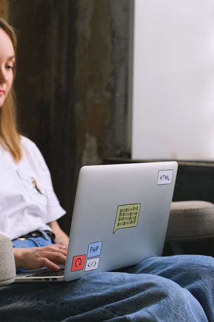 A person with long, light brown hair, wearing a white shirt and blue jeans, is seated outdoors with their legs crossed. They are working on a silver laptop placed on their lap, which features various